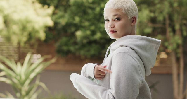Young woman with short blonde hair pointing at her grey hoodie while standing outside in a lush, green garden. Ideal for fashion editorials, advertising casual wear, and lifestyle blogs aiming to capture modern, casual styles.