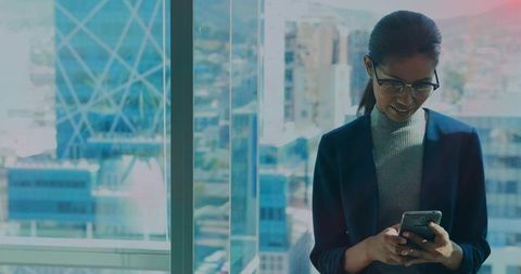 Professional woman standing by office window using smartphone with city skyline view