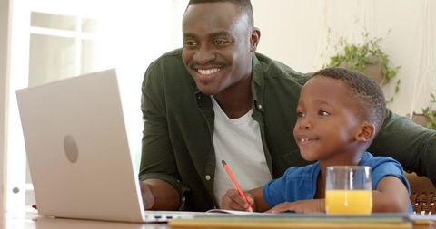 African american father helping son with homework using laptop and notebook, smiling