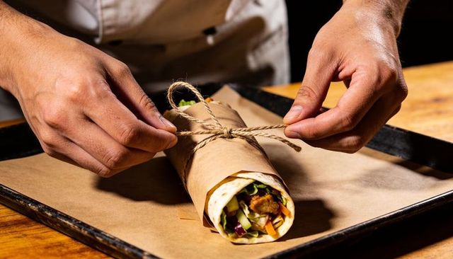 Chef tying kraft-wrapped burrito with twine on tray, hands preparing gourmet wrap