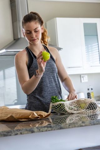 Young woman unpacking groceries holding green apple checking ripeness in modern kitchen