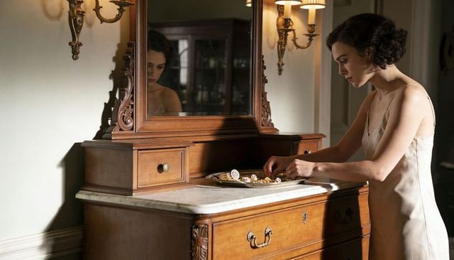 Light-skinned woman in pale silk slip arranging jewelry on marble-top antique vanity mirror