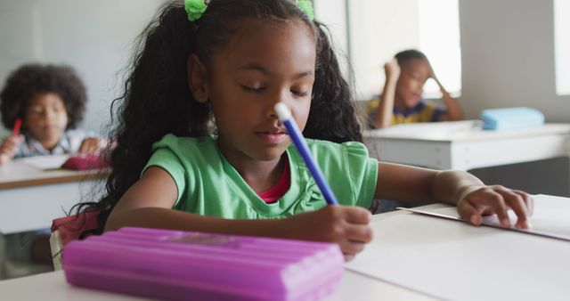 Young girl with curly hair writing intently while sitting at a desk in a classroom. Other students are visible in the background. Useful for educational content, school advertisements, learning programs, and child development resources.