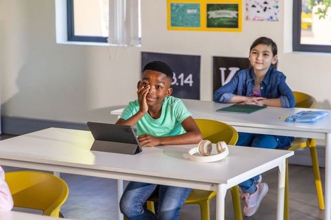 Young african american boy using tablet in bright classroom with attentive asian classmate