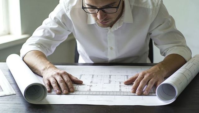 Architect studying detailed architectural blueprints with scale ruler and rolled plans at desk