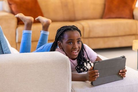 African american girl using tablet while lying on ottoman at home enjoying learning