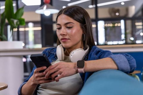 Young woman relaxing on blue couch scrolling smartphone in modern coworking lounge
