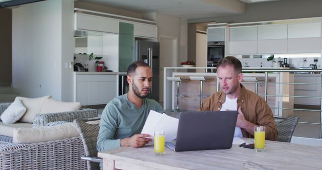 Two men sitting at a wooden table in a modern home, reviewing documents and working together on a laptop. Both have glasses of juice, suggesting a casual, friendly atmosphere. Ideal for topics related to remote work, teamwork, home office setups, collaboration, and modern lifestyle themes.