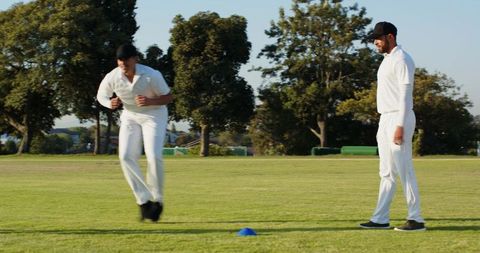 Cricket players training with coach bounding over cone on sunny grass practice field drill