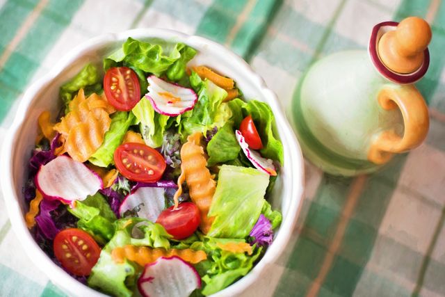 Fresh vegetable salad with lettuce, cherry tomatoes, radishes, and crinkle-cut carrots in ceramic bowl on green checkered tablecloth beside ceramic bottle for vinaigrette. Perfect for promoting healthy eating, vegetarian and vegan recipes, organic farming, summer meals, and diet plans.