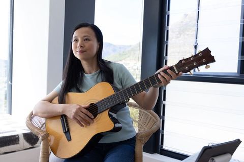 Mature asian woman playing acoustic guitar in sunlit rattan chair by window with tablet
