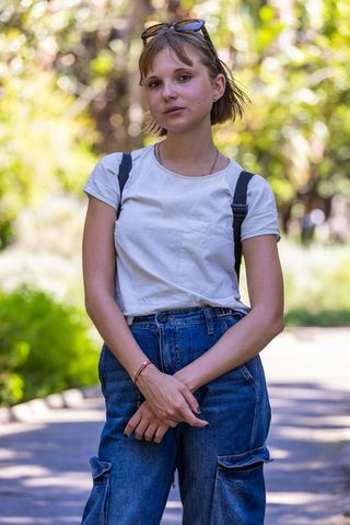 Standing on sunlit park path wearing white t-shirt, denim jeans, sunglasses and backpack