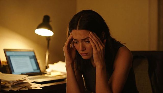 Late-night overwork: woman holding head at desk with laptop and scattered papers