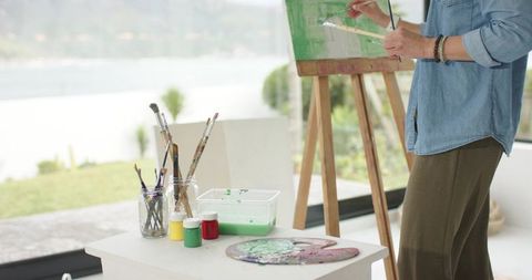 Female artist painting lakeside canvas at sunlit studio easel with brushes and palette