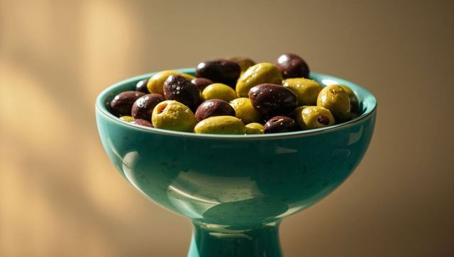 Teal pedestal bowl overflowing with glossy green and purple olives in warm studio light