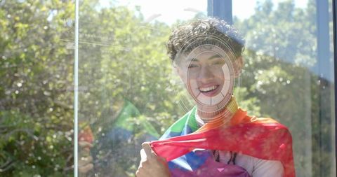 Smiling man wrapped in rainbow flag outdoors with circular reflection and sunlight