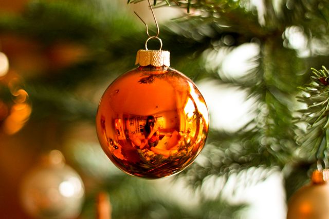 Golden Christmas ornament hanging on an evergreen branch. This decorative sphere reflects a cozy indoor setting and adds festive charm to the holiday decor. Perfect for use in holiday greeting cards, decorations, festive advertising, or social media posts celebrating the Christmas season.