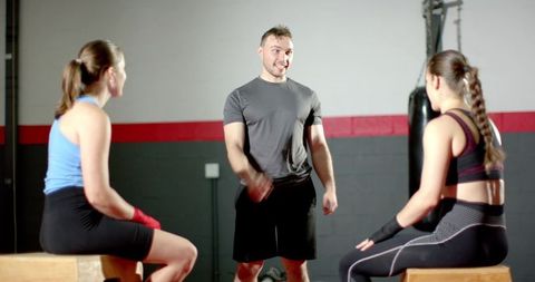Male fitness coach guiding two female trainees on plyo boxes by heavy bag in industrial gym