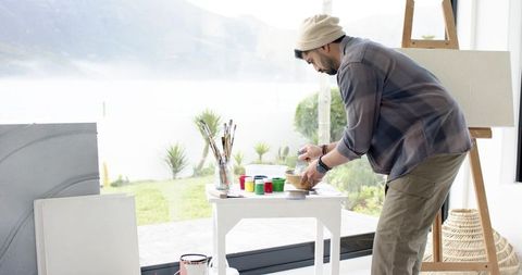 Indian young artist arranging paints and brushes by lakeside window in daylight studio