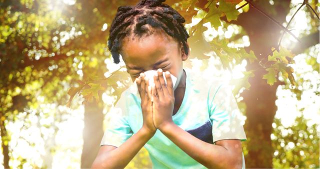 Young child sneezing into a tissue while standing in a sunlit forest. Sunlight filters through the green leaves, evoking a sense of nature and outdoor activities. This image can be used in articles about seasonal allergies, children's health, nature experiences, environmental education, and outdoor life.