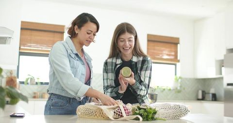 Diverse women unpacking reusable grocery bags inspecting jar and fresh produce in kitchen