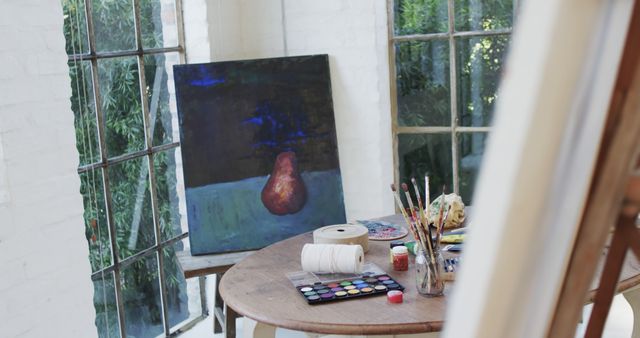 Art studio featuring an easel with a painting of a pear, wooden table filled with painting supplies including brushes, paints, and a palette. Natural light streams in through the large windows. Suitable for use in content related to art creation, artist's life, and creative workspaces.