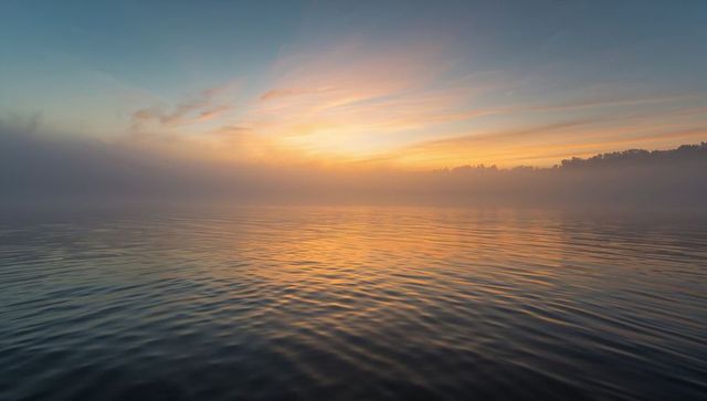 Misty sunrise reflecting warm pastel light on rippled lake water with distant treeline