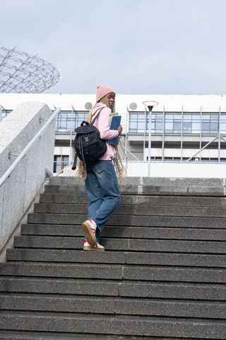 Young african american student climbing urban stairs carrying backpack and books