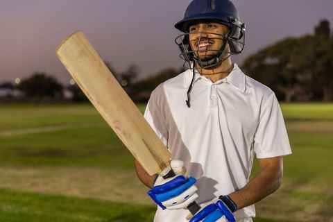 Indian cricketer smiling holding bat at dusk on grass cricket field wearing helmet and gloves