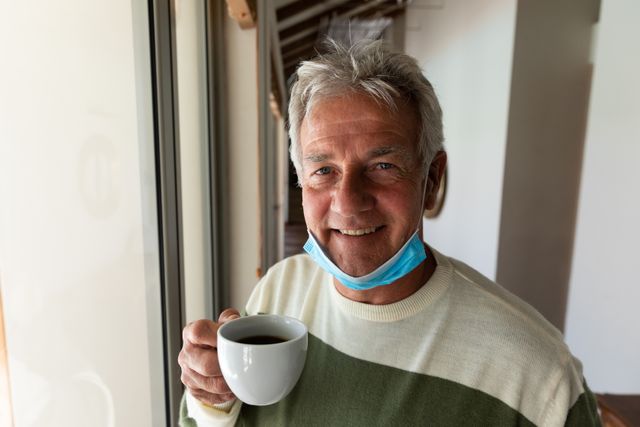 Senior man enjoying a cup of coffee at home with a lowered face mask. Ideal for use in articles about retirement lifestyle, health and safety during the pandemic, morning routines, and senior well-being.