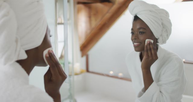 Young African American woman applying skincare product with cotton pad, smiling and looking in mirror. Ambiance feels calm and relaxing, highlighting wellness and self-care. Ideal for promoting skincare products, spa treatments, beauty routines, and wellness campaigns.