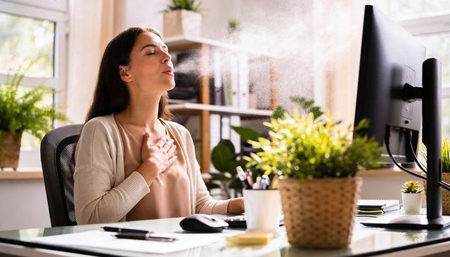 Woman exhaling and clutching chest at bright plant-filled office desk, wellness and relief