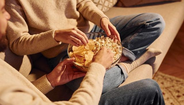 Couple sharing popcorn and potato chips on cozy sofa in sweaters and jeans