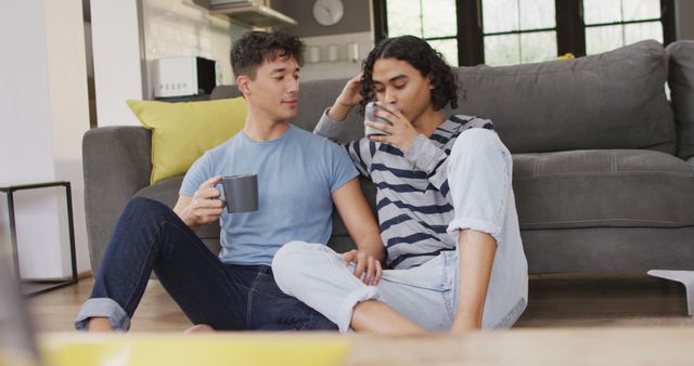 Happy diverse male couple sitting on floor and drinking coffee in living room. Spending quality time at home.