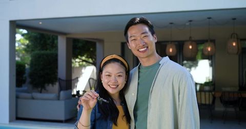 Asian couple celebrating new home by modern backyard pool holding single house key