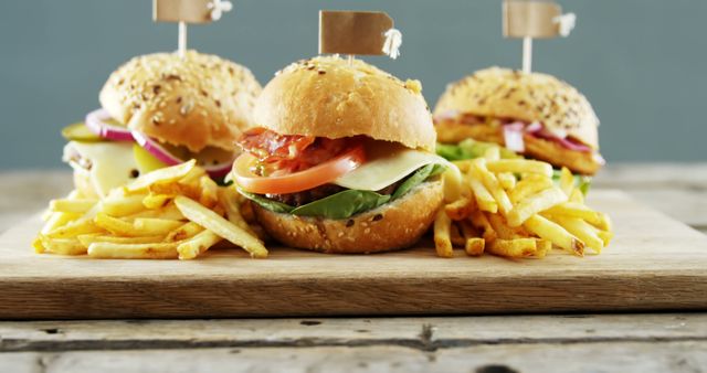 Three gourmet burgers with fresh vegetables, including lettuce, tomato, and onion, are served with crispy French fries on a wooden board. Ideal for use in food industry marketing materials, restaurant menus, culinary blogs, or advertisements for fast food and American cuisine.