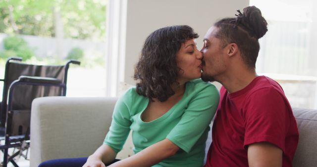 Happy biracial couple sitting on couch kissing in living room, with wheelchair in background - Download Free Stock Photos Pikwizard.com