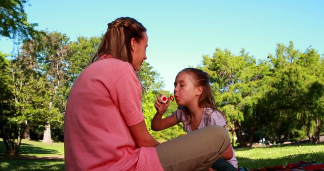 Mother and daughter spending quality time in a park on a sunny day. Daughter blowing bubbles while mother watches, both sitting on blanket. Ideal for promoting family bonding, outdoor activities, summer fun, or parenting content.