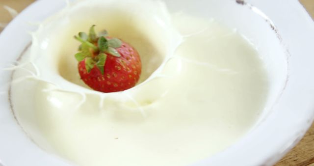 A fresh strawberry falling into cream creates a splash in a porcelain bowl. Useful for food blogs, restaurant menus, advertisements for dairy products, or depicting fresh, wholesome ingredients in culinary contexts.