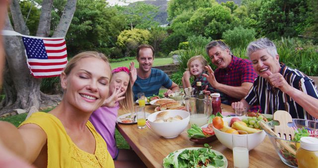 Smiling caucasian woman taking selfie with three generation family having celebration meal in garden. three generation family celebrating independence day eating outdoors together.
