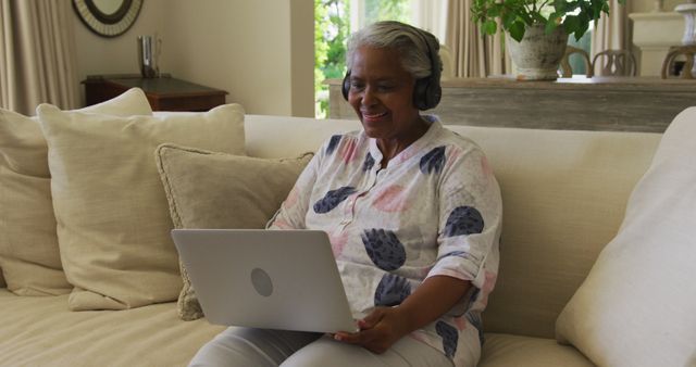African american senior woman wearing headphones smiling while having a image call on laptop at home. retirement senior lifestyle living concept