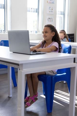 Smiling schoolgirl using laptop at desk in sunlit classroom with artwork and windows