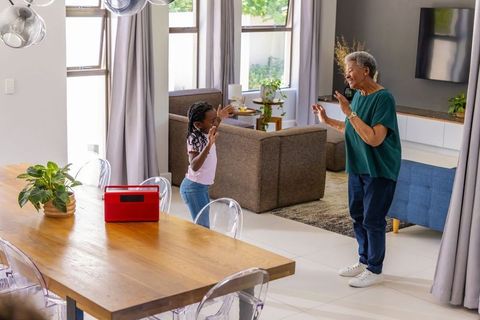 African american grandmother and granddaughter dancing in sunny living room with red speaker