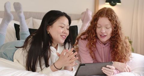 Diverse female friends relaxing on bed browsing tablet together in cozy loungewear