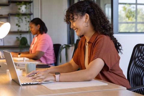 Hispanic woman typing on laptop in modern coworking office, smiling coworker in background