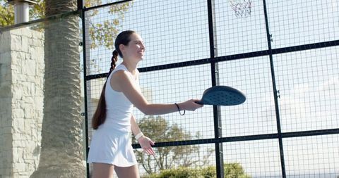 Young woman playing pickleball on waterfront fenced court in white athletic dress