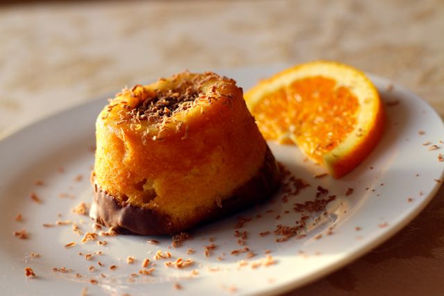 Orange cake with chocolate topping garnished with chocolate shavings served with an orange slice on a white plate. Ideal for illustrating homemade desserts, gourmet treats, and food blogs. Perfect for use in recipe websites, baking tutorials, or social media posts promoting sweet treats.