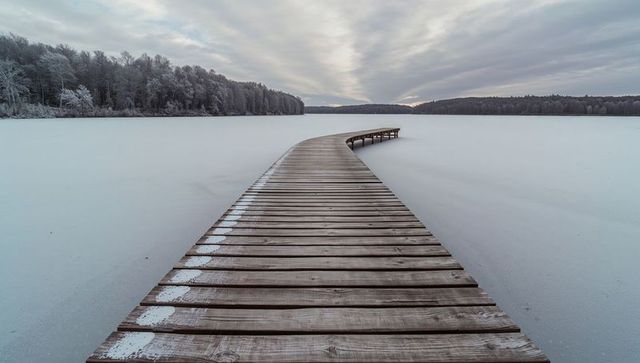 Curving wooden pier leading across frozen lake toward frosted treeline under winter sky