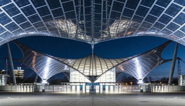 Illuminated steel-and-glass tensile canopy framing central dome over urban plaza at night