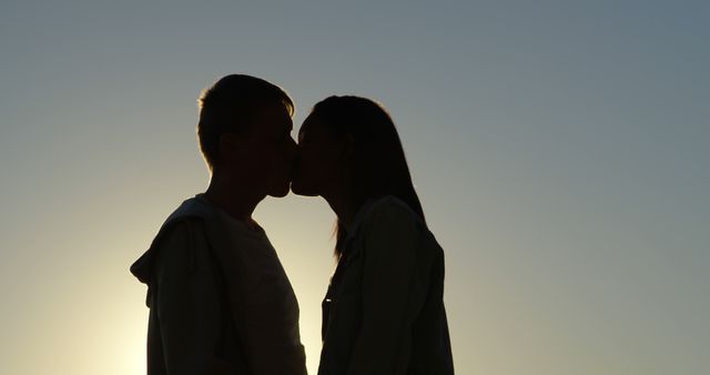 Silhouetted against a serene sky, a young Caucasian couple shares a romantic kiss, with copy space. Their affectionate moment captures the essence of love and intimacy at dusk.